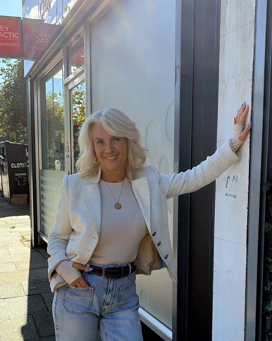 Laurie Traquair smiling, one hand on the wall, in a cream blazer and jeans
